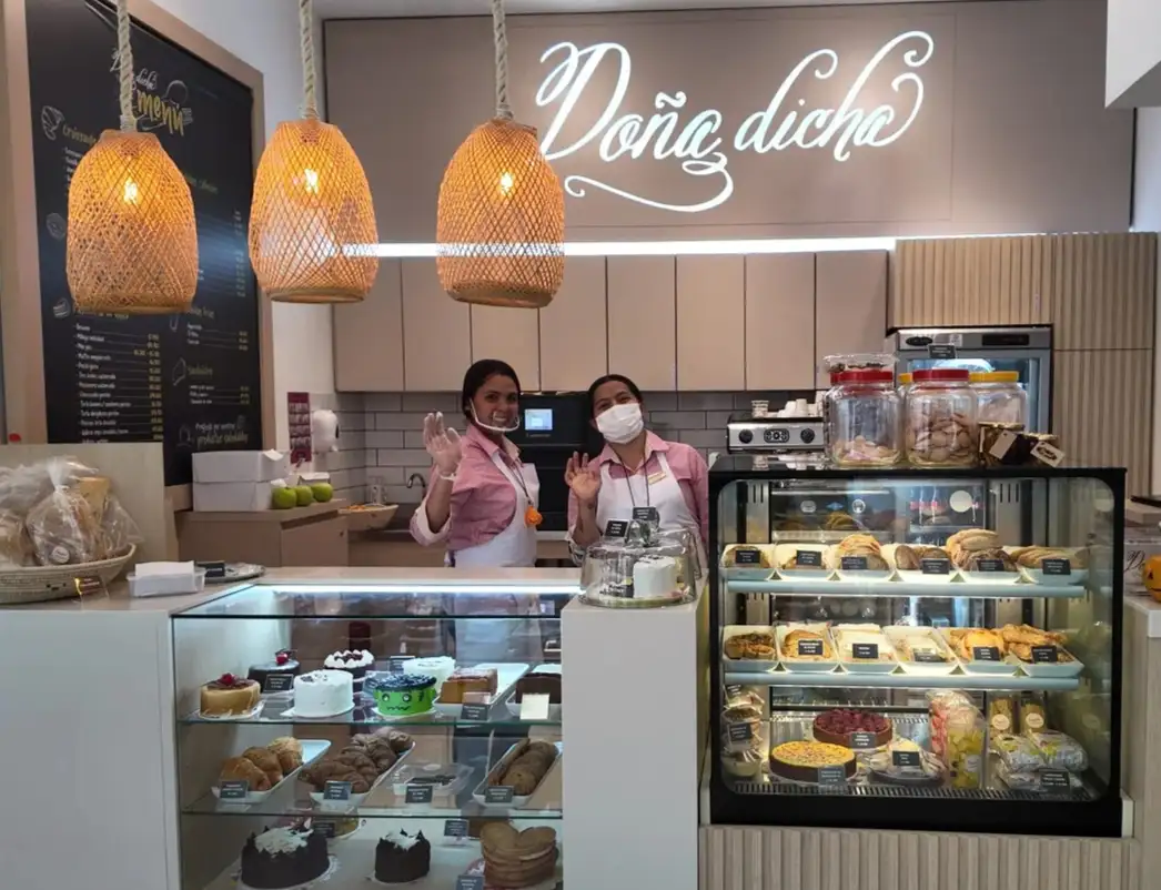 Dos colaboradoras del equipo de la pastelería Doña Dicha en Bogotá saludando detrás del mostrador con vitrinas de tortas y postres.