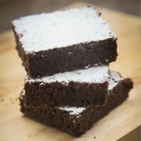 Torre de tres brownies artesanales melcochudos espolvoreados con azúcar glass sobre tabla.