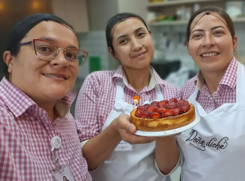 Trabaja con Nosotros - Equipo Doña Dicha Bogotá Tres colaboradoras del equipo de Doña Dicha sonriendo mientras sostienen un pastel de fresa.