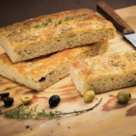 Porciones de focaccia artesanal con hierbas finas y aceitunas sobre una tabla de madera.